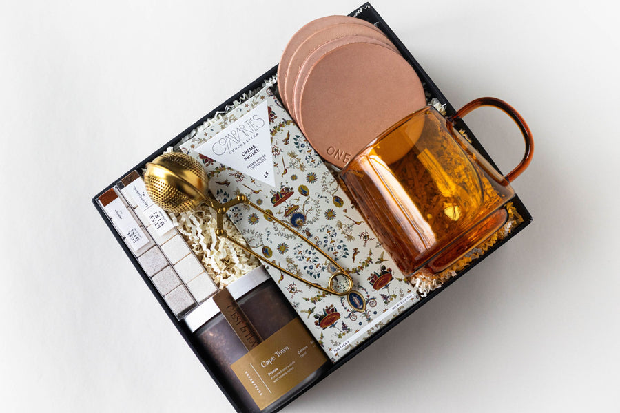 gift box shown open with lid - includes leather coasters, 2 packs of mini sugar cubes, amber-colored glass mug, creme brulee chocolate bar, jar looseleaf tea and gold tea strainer with handle.