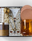 gift box shown open with lid - includes leather coasters, 2 packs of mini sugar cubes, amber-colored glass mug, creme brulee chocolate bar, jar looseleaf tea and gold tea strainer with handle.