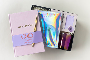 purple gift box for her containing a linen journal with the words 'vibras bonitas' debossed on the front, matching gold pen, irredescent colored facial mask and candle, and a purple satin pillowcase arranged in a black gift box.