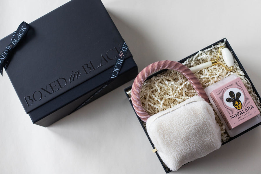 pink themed self care gift set includes an unwind kit in plush pouch, pink velvet headband, white and gold facial roller, pink cactus bar soap and concrete soap dish in a black keepsake box shown with lid.