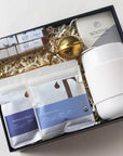 Complete tea set shown with box and lid - includes 2 tea packets (Manhattan Black and Charles Grey), 2 packs of luxe mini sugar cubes in London Fog and Vanilla, a lavender bath bomb, cream colored portable mug and gold tea strainer.