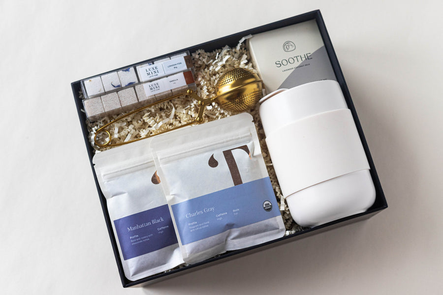 Complete tea set shown with box and lid - includes 2 tea packets (Manhattan Black and Charles Grey), 2 packs of luxe mini sugar cubes in London Fog and Vanilla, a lavender bath bomb, cream colored portable mug and gold tea strainer.
