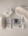 contents of the unwind self care kit - includes plush pouch, satin scrunchie, shower steamers, facial cleanser, lip balm, plush sleep mask and white and gold facial roller.