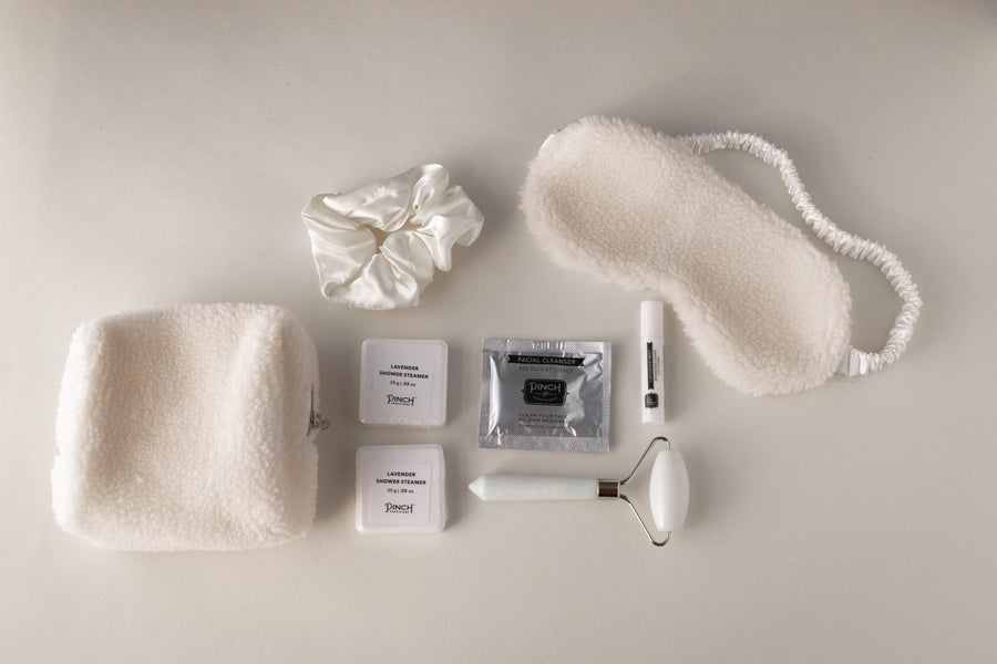 contents of the unwind self care kit - includes plush pouch, satin scrunchie, shower steamers, facial cleanser, lip balm, plush sleep mask and white and gold facial roller.